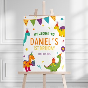 May include: A welcome sign for a 1st birthday party with a dinosaur theme. The sign is white with colorful dinosaurs, stars, and confetti. The text reads "Welcome to Daniel's 1st Birthday 20th July 2025".