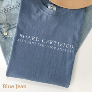 May include: A blue t-shirt with the words "BOARD CERTIFIED ASSISTANT BEHAVIOR ANALYST" printed in white. The shirt is folded on top of a pair of blue jeans. A gold necklace and a tan hat are also visible.