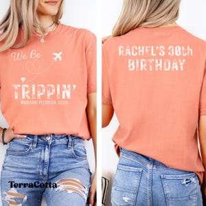 May include: A terracotta-colored t-shirt with the text "We Be Trippin'" and a graphic of a plane and heart. The text "Miami Florida 2026" is below. The back of the shirt says "Rachel's 30th Birthday".