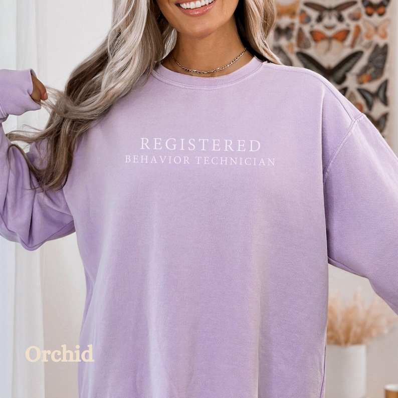 Pode incluir: Uma camisa de moletom lil&aacute;s com o texto "REGISTERED BEHAVIOR TECHNICIAN" impresso em branco.