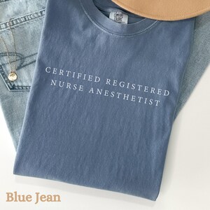 May include: A blue Comfort Colors t-shirt with the text "CERTIFIED REGISTERED NURSE ANESTHETIST" in white. The shirt is folded on top of a pair of blue jeans, with a gold necklace and a tan hat visible.