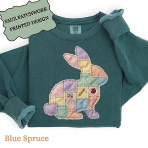 May include: Blue spruce sweatshirt with a faux patchwork rabbit design. The rabbit is made of pastel-colored squares and features a pencil, apple, and books. The text "FAUX PATCHWORK PRINTED DESIGN" is at the top.