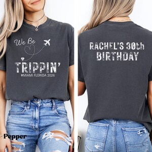May include: Dark gray t-shirt with white text and graphics. The front of the shirt says "We Be Trippin'" with a heart and airplane graphic, and "Miami Florida 2026". The back says "Rachel's 30th Birthday".