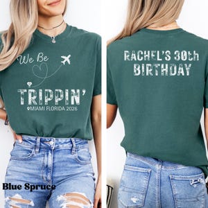 May include: Teal-colored t-shirt with the text "We Be Trippin' Miami Florida 2026" and a graphic of a plane and heart on the front. The back of the shirt says "Rachel's 30th Birthday" in white letters. The shirt is paired with blue jeans.