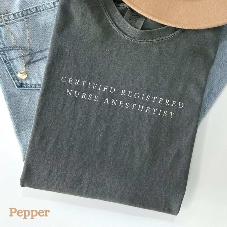 May include: A dark gray t-shirt with the text "CERTIFIED REGISTERED NURSE ANESTHETIST" in white. The shirt is folded on top of a pair of light blue jeans. A gold necklace and a tan hat are also visible.