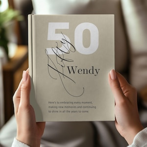 May include: A beige book with the number "50" in white, the name "Wendy" in black, and cursive text. The book's cover also includes the text "Here's to embracing every moment, making new memories and continuing to shine in all the years to come."