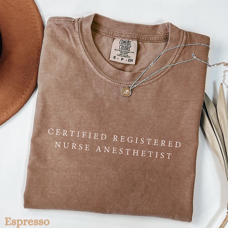 May include: Espresso-colored t-shirt with the text "CERTIFIED REGISTERED NURSE ANESTHETIST" in white. A silver necklace with a pendant rests on the shirt. The shirt is folded and displayed with a brown hat and decorative elements.