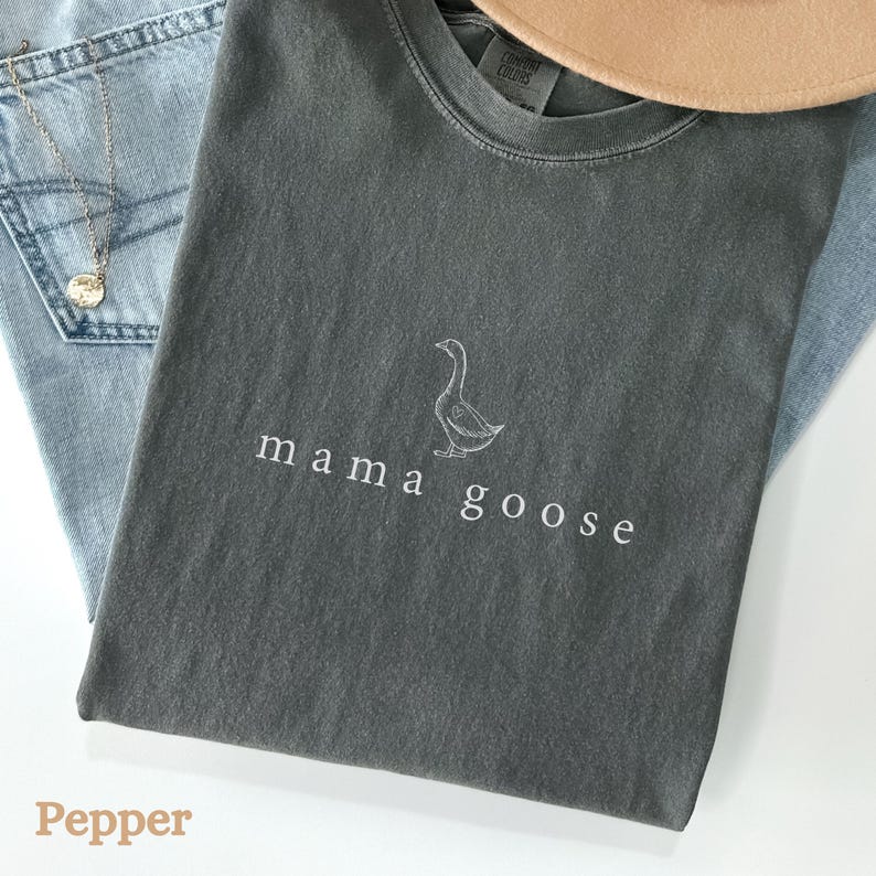以下が含まれることがあります： 暗いグレーのTシャツに、白い刺繍のガチョウと「mama goose」の文字が刺繍されています。Tシャツは青いデニムの上に平らに置かれています。