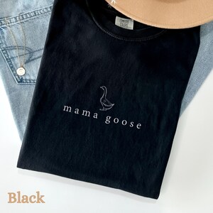 以下が含まれることがあります： 前面にガチョウの白いアウトラインと「mama goose」という文字がプリントされた黒いTシャツです。