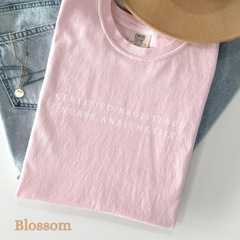 May include: A light pink t-shirt with the text "CERTIFIED REGISTERED NURSE ANESTHETIST" in white. The shirt is folded on top of blue jeans, with a gold necklace and a tan hat visible.