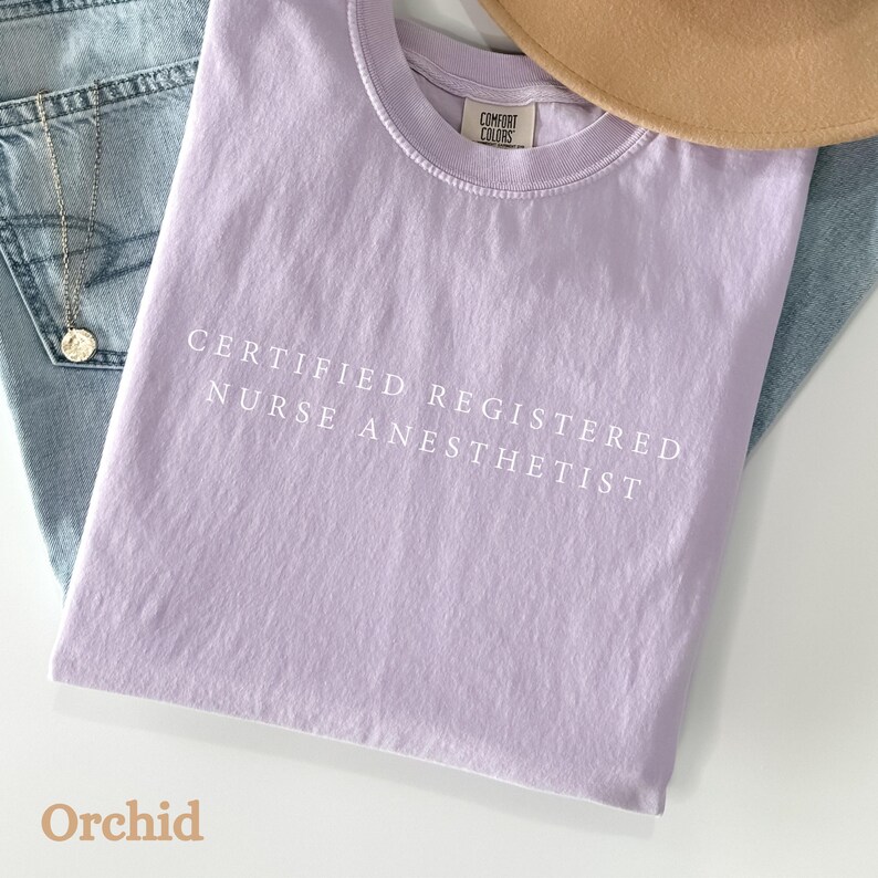 May include: A light purple t-shirt with the text "CERTIFIED REGISTERED NURSE ANESTHETIST" in white. The shirt is folded and rests on a pair of blue jeans. A gold necklace and a tan hat are also visible.