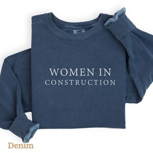 Op de afbeelding: Een donkerblauw sweatshirt met de tekst "WOMEN IN CONSTRUCTION" in wit gedrukt op de voorkant.