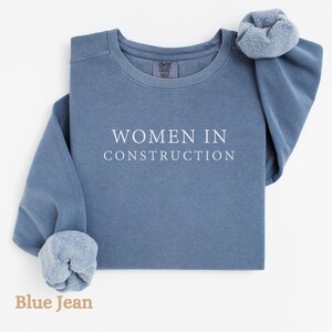 Op de afbeelding: Een blauw jeans sweatshirt met de tekst "WOMEN IN CONSTRUCTION" in wit gedrukt op de voorkant.