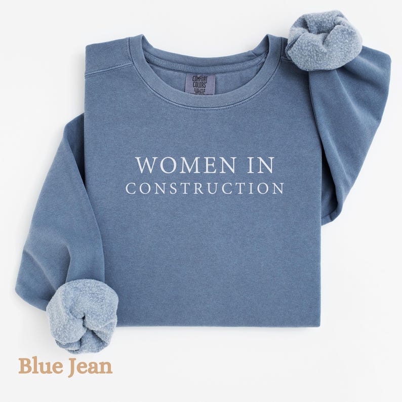 Op de afbeelding: Een blauw jeans sweatshirt met de tekst "WOMEN IN CONSTRUCTION" in wit gedrukt op de voorkant.