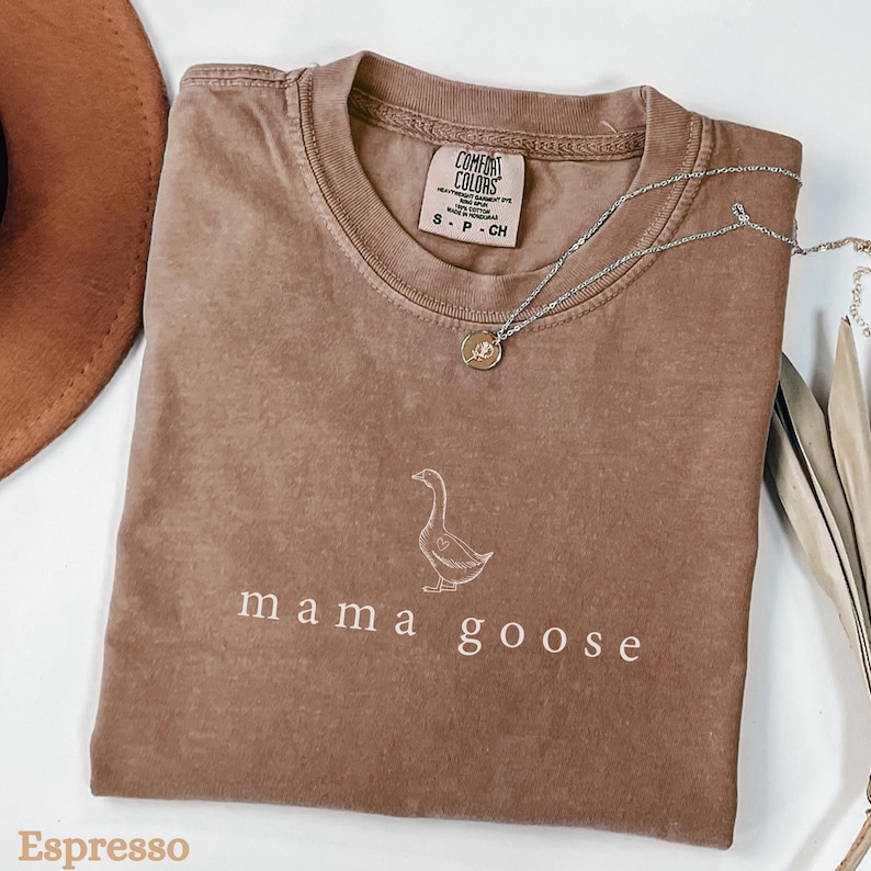 以下が含まれることがあります： 白い輪郭のガチョウと「mama goose」という文字が書かれた茶色のTシャツです。