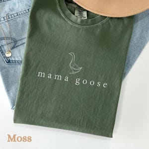 以下が含まれることがあります： モスグリーンのTシャツに、白いアウトラインのガチョウと「mama goose」の文字が前面にプリントされています。