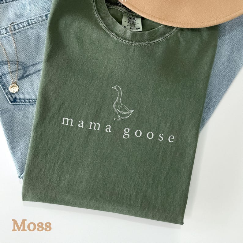 以下が含まれることがあります： モスグリーンのTシャツに、白いアウトラインのガチョウと「mama goose」の文字が前面にプリントされています。