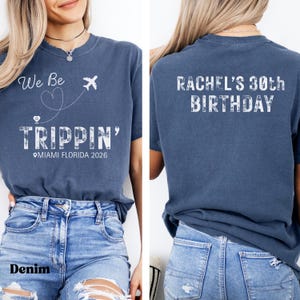 May include: Denim blue t-shirt with white text and graphics. The front of the shirt says "We Be Trippin'" with a heart, airplane, and the location "Miami Florida 2026." The back says "Rachel's 30th Birthday."