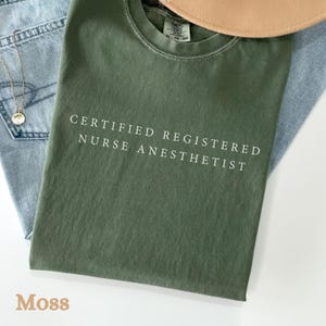 May include: A moss green t-shirt with the text "CERTIFIED REGISTERED NURSE ANESTHETIST" in white. The shirt is folded on top of a pair of blue jeans and a tan hat. A gold necklace with a pendant is visible.