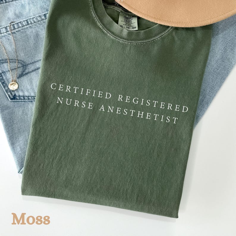 May include: A moss green t-shirt with the text "CERTIFIED REGISTERED NURSE ANESTHETIST" in white. The shirt is folded on top of a pair of blue jeans and a tan hat. A gold necklace with a pendant is visible.