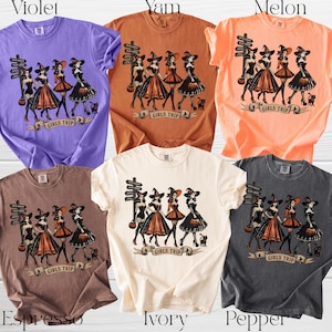 May include: A collection of six short-sleeved t-shirts in violet, yam, melon, espresso, ivory, and pepper colors. Each shirt features a graphic print of four witches standing near a signpost with the text Girls Trip and a small black cat.
