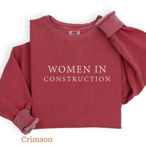 Op de afbeelding: Een crimsonrode trui met de tekst "WOMEN IN CONSTRUCTION" in wit.
