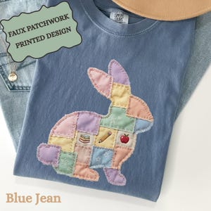 May include: A blue jean-colored t-shirt with a faux patchwork rabbit design. The rabbit is made of pastel-colored squares and features a pencil, apple, and book graphic. The shirt also has the text "FAUX PATCHWORK PRINTED DESIGN".