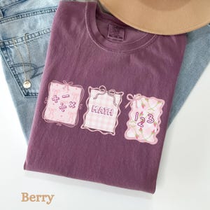 May include: Berry-colored t-shirt with three pink framed designs. The designs include math symbols, the word "MATH", and the numbers "123". The shirt is paired with blue jeans and a tan hat.