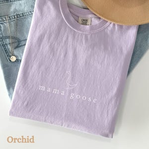 以下が含まれることがあります： 薄い紫色のTシャツに「mama goose」の文字と、白いアウトラインで描かれた小さなガチョウがプリントされています。このTシャツはComfort Colors製で、色はOrchidです。