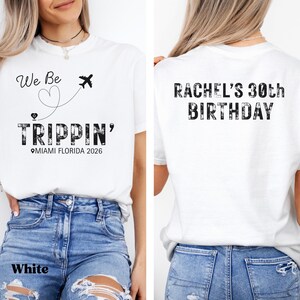 May include: White t-shirt with the text "We Be Trippin' Miami Florida 2026" on the front, and "Rachel's 30th Birthday" on the back. The front design includes a heart, airplane, and location pin. The shirt is paired with blue jeans.