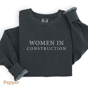 Op de afbeelding: Een donkergrijs sweatshirt met de tekst "WOMEN IN CONSTRUCTION" in wit gedrukt op de voorkant.