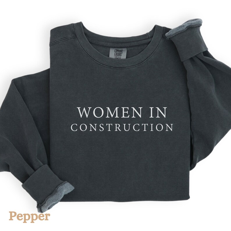Op de afbeelding: Een donkergrijs sweatshirt met de tekst "WOMEN IN CONSTRUCTION" in wit gedrukt op de voorkant.