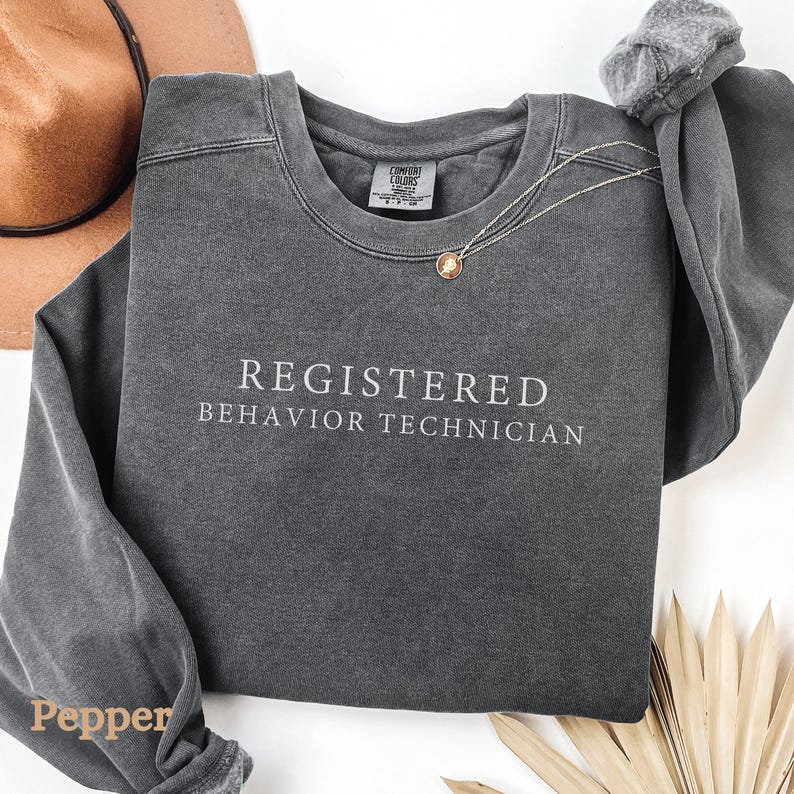 Pode incluir: Uma camisa de moletom cinza escuro com o texto "REGISTERED BEHAVIOR TECHNICIAN" impresso em branco na frente.