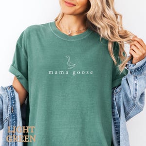 以下が含まれることがあります： 薄い緑色のTシャツに、白いアウトラインのガチョウと「mama goose」という文字が前面にプリントされています。