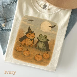 May include: Ivory-colored t-shirt featuring a Halloween-themed illustration. The design includes two frogs dressed in witch and vampire costumes, pumpkins, bats, and a full moon. The shirt is made of a soft, comfortable material.