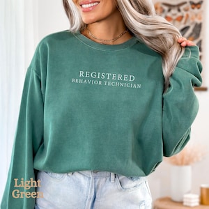 Pode incluir: Uma camisa de moletom verde claro com o texto "REGISTERED BEHAVIOR TECHNICIAN" impresso em branco.