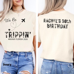 May include: Ivory-colored t-shirt with the text "We Be Trippin' Miami Florida 2026" on the front, featuring a heart and airplane graphic. The back of the shirt says "Rachel's 30th Birthday".