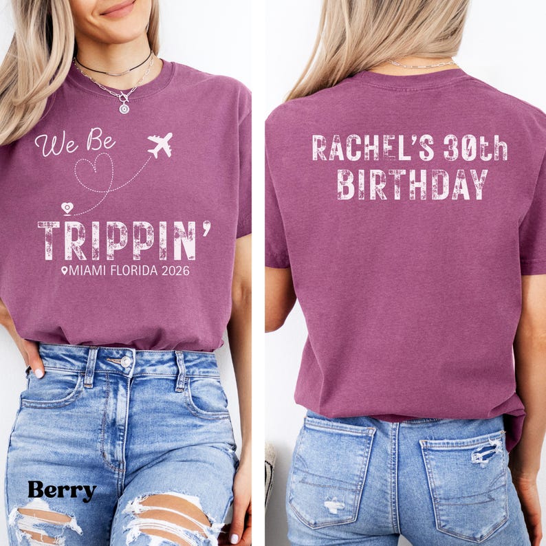 May include: Berry-colored t-shirt with the text "We Be Trippin' Miami Florida 2026" on the front. The back of the shirt says "Rachel's 30th Birthday" in white lettering. The shirt is paired with blue denim jeans.