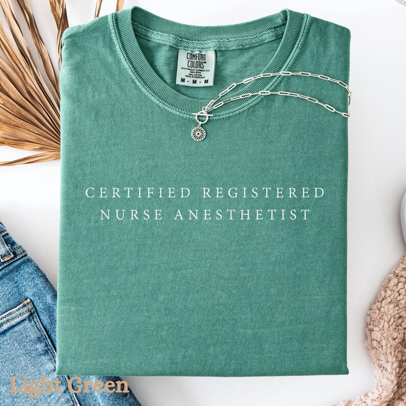 May include: A light green t-shirt with the text "CERTIFIED REGISTERED NURSE ANESTHETIST" in white. A silver necklace with a pendant is draped across the shirt. The shirt is folded neatly, with the brand tag visible.
