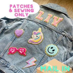 May include: A light blue denim jacket adorned with colorful patches. Patches include a heart, a bow, a lightning bolt, a smiling face, a unicorn, and the name "LIV". The patches are in pink, yellow, blue, and gold.