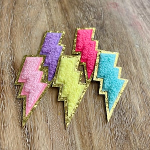 May include: A collection of colorful lightning bolt patches. Each patch features a textured, felt-like design in shades of pink, purple, yellow, and turquoise, outlined with gold glitter. These patches are ideal for adding a pop of color to clothing or accessories.