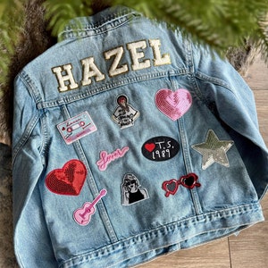 May include: Light blue denim jacket with the name "HAZEL" embroidered on the back. The jacket is decorated with various patches, including hearts, a guitar, sunglasses, and the text "Lover" and "T.S. 1989".