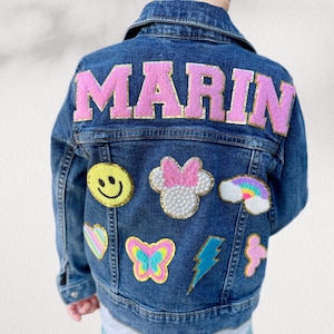 May include: A denim jacket with the name "MARIN" in pink, gold-trimmed letters on the back. The jacket features various colorful patches, including a smiley face, a Minnie Mouse head, a rainbow, a heart, a butterfly, a lightning bolt, and a flower.