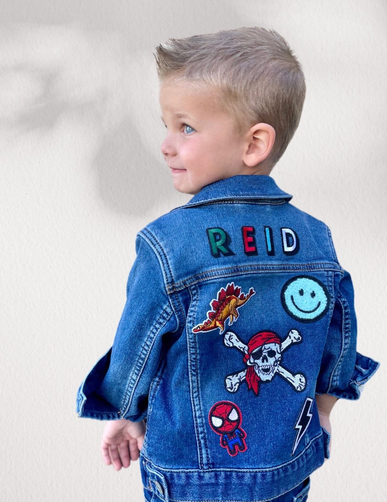 May include: A blue denim jacket with patches, including a dinosaur, a skull and crossbones, a smiley face, a spider-man, and a lightning bolt. The name "REID" is embroidered on the back in colorful letters.