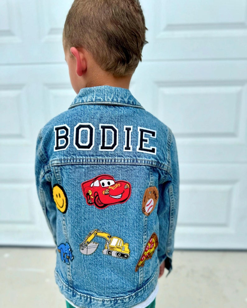 May include: A light blue denim jacket with the name "BODIE" in white letters with a black outline. The jacket is decorated with patches including a red race car, a yellow excavator, a baseball, a slice of pizza, a smiley face, and a blue dinosaur.