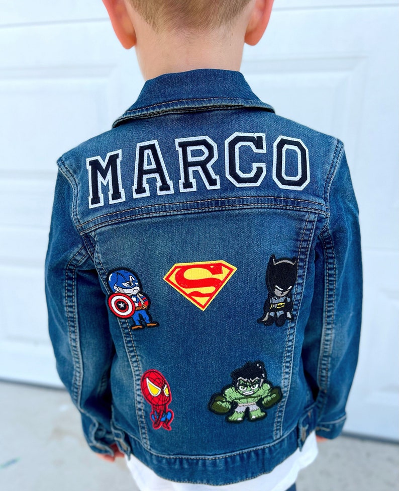 May include: A denim jacket with the name "MARCO" in white block letters with a black outline. The jacket is decorated with superhero patches, including Captain America, Superman, Batman, Spider-Man, and the Hulk.