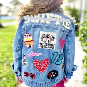May include: A light blue denim jacket with various patches, including a red heart, the number 13, and the text "TAYLOR SWIFT THE ERAS TOUR." The jacket has gold lettering on the back and is worn over a pink sequined dress.