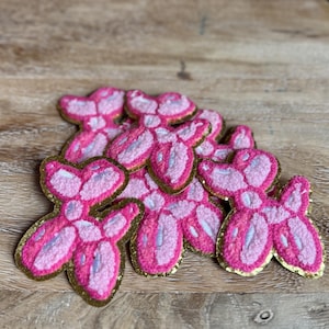 May include: A collection of pink balloon dog patches featuring a textured surface with light pink and dark pink thread details, accented by white highlights and a shimmering gold metallic border outlining each individual dog shape.