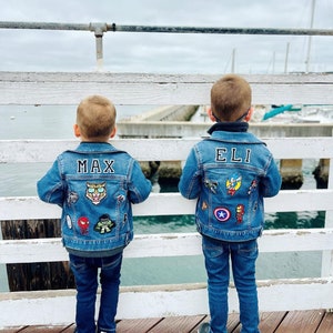 May include: Two children wearing denim jackets with patches and names on the back. The jacket on the left says "MAX" and has a tiger patch. The jacket on the right says "ELI" and has a Captain America patch. They are standing on a wooden pier.