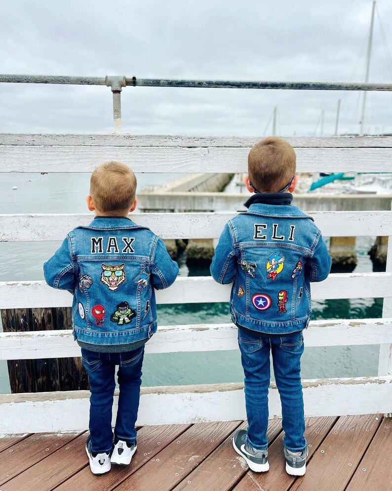 May include: Two children wearing denim jackets with patches and names on the back. The jacket on the left says "MAX" and has a tiger patch. The jacket on the right says "ELI" and has a Captain America patch. They are standing on a wooden pier.